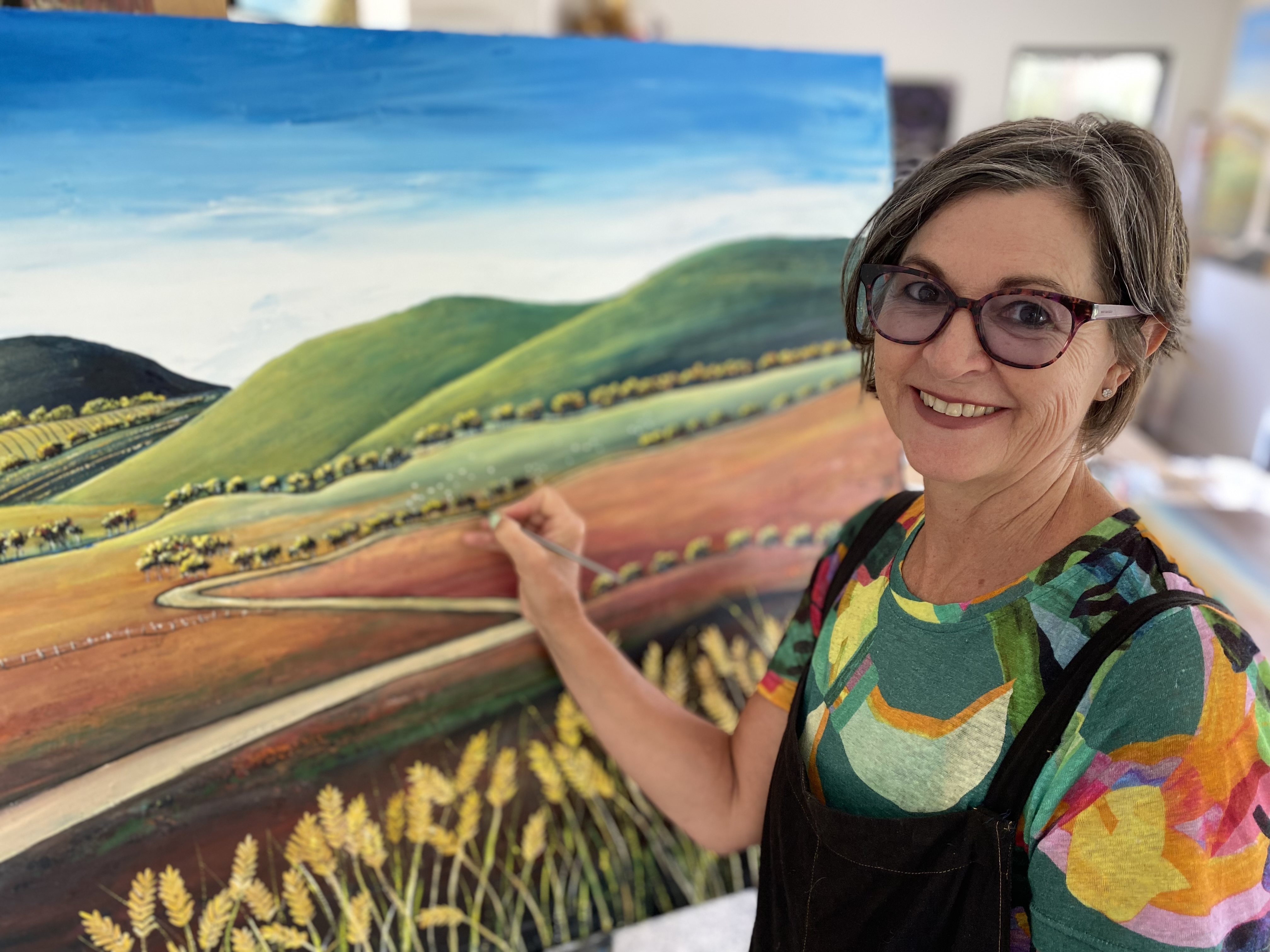 Corrie Young holds a paint brush next to one of her landscape paintings