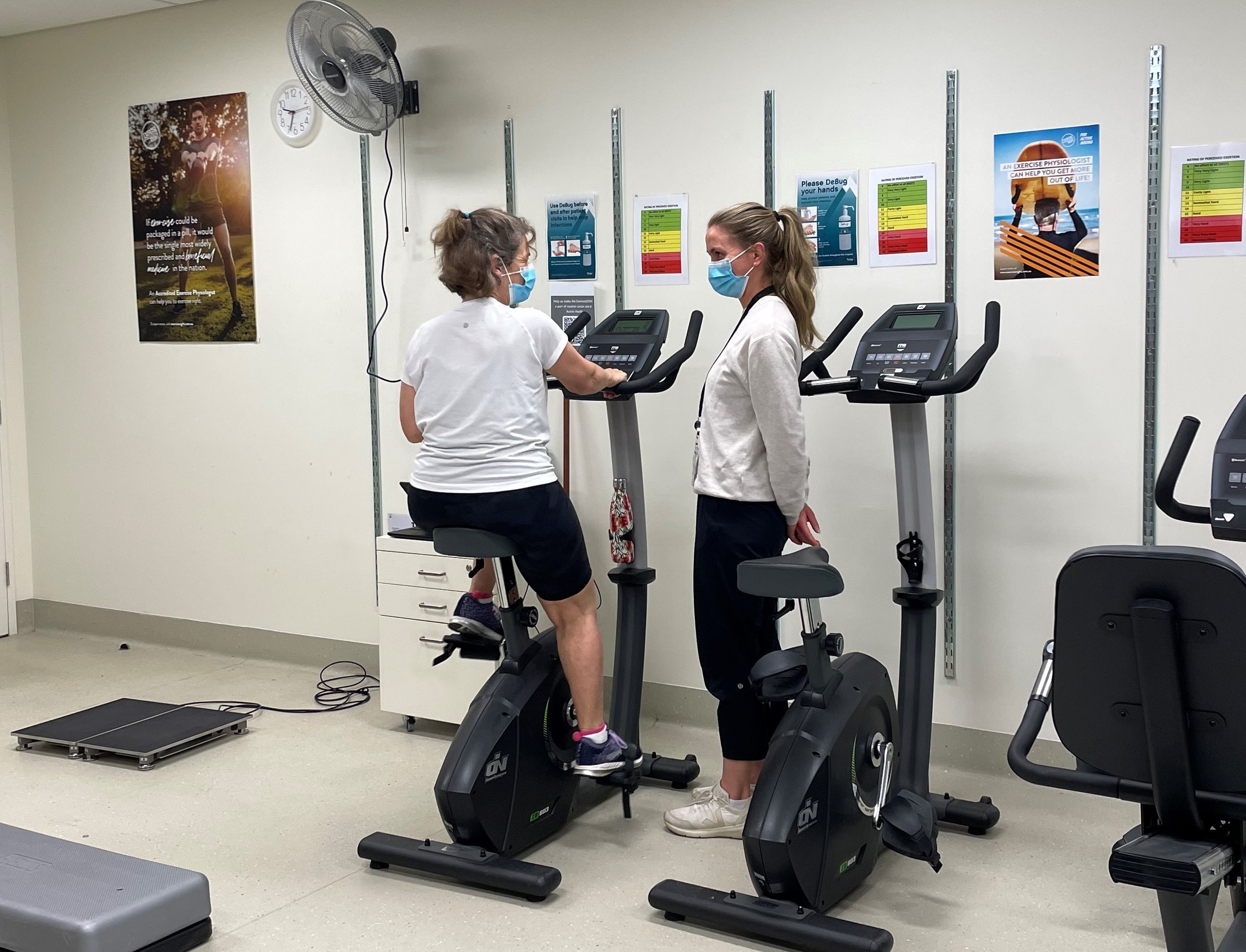 Grace stands next to a patient on an exercise bike