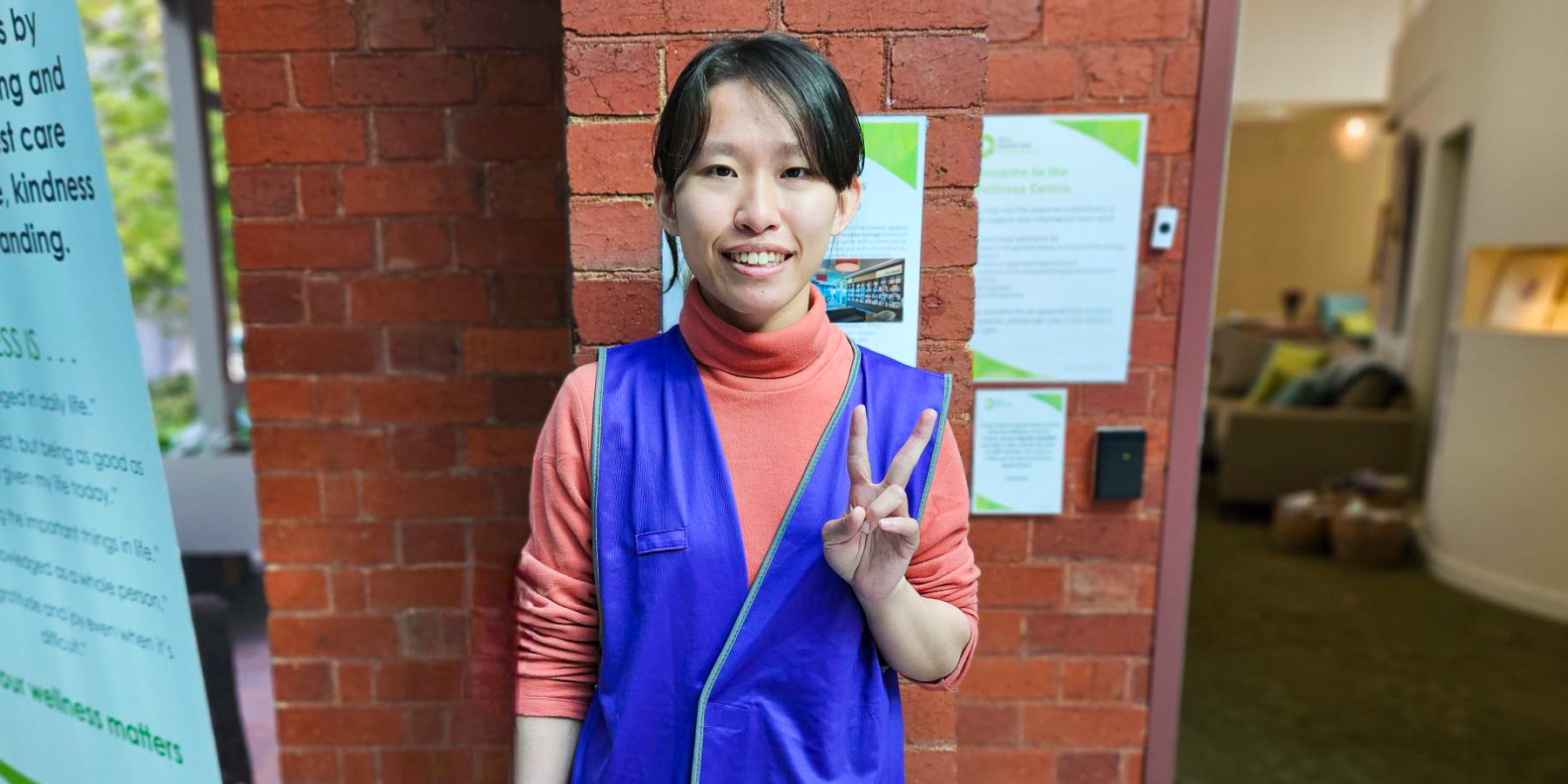 Picture of ONJ Centre volunteer Lynn Lin smiling at camera, holding up peace sign. She wears a purple volunteer vest.
